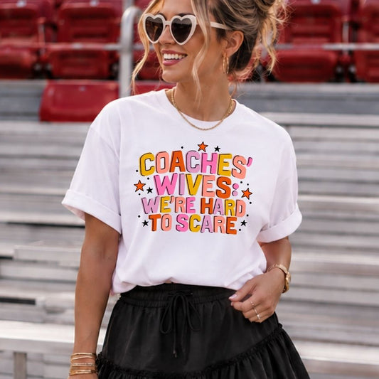 A woman wearing a white graphic t-shirt with the text 'COACHES' WIVES WE'RE HARD TO SCARE' in colorful letters, paired with a black ruffled mini skirt, standing in stadium seating.