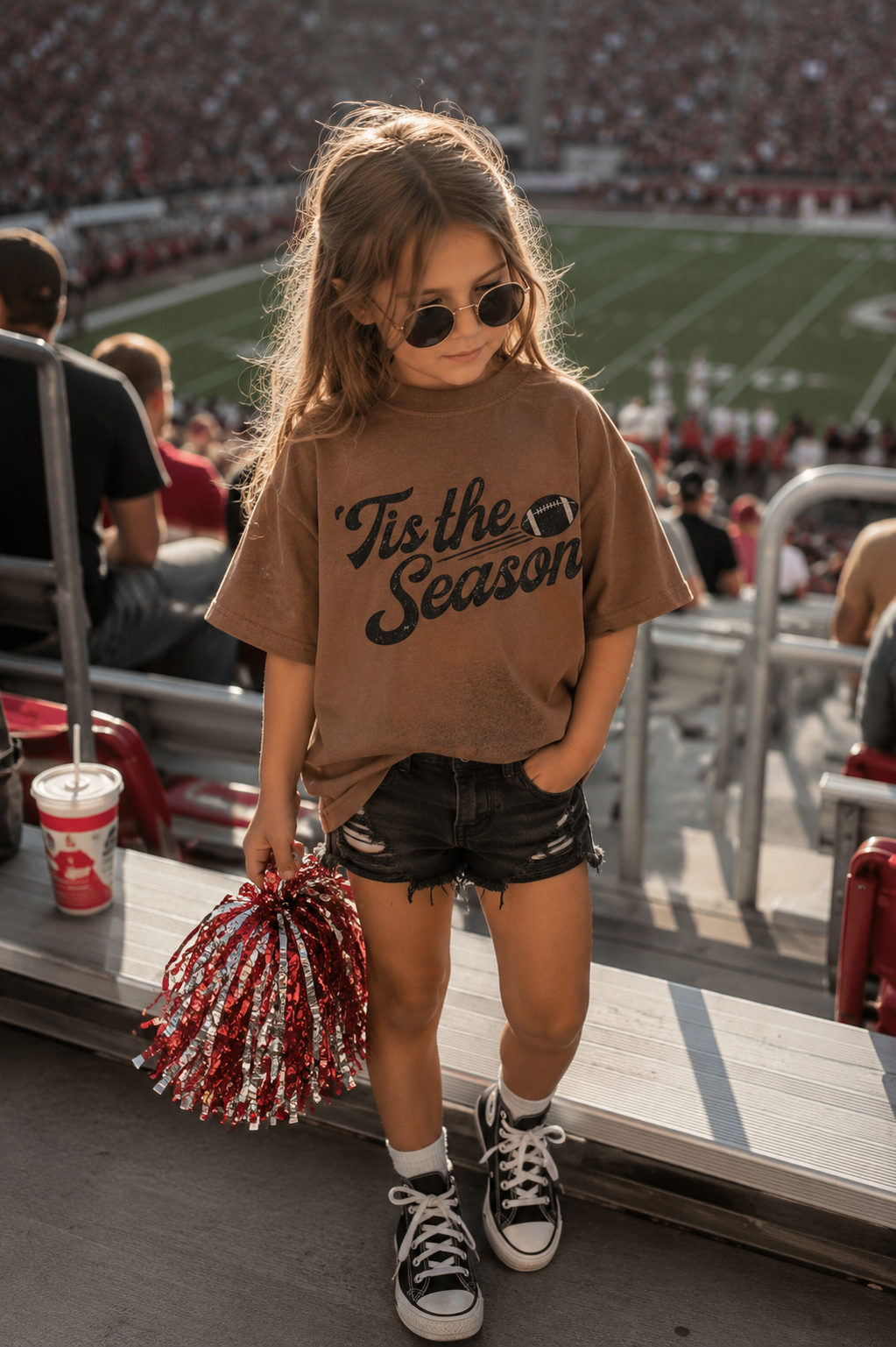 A young girl stands in a stadium wearing a brown graphic t-shirt that says 'Tis the Season' in black script, black denim shorts, black sneakers, and sunglasses, holding red and white pom-poms.
