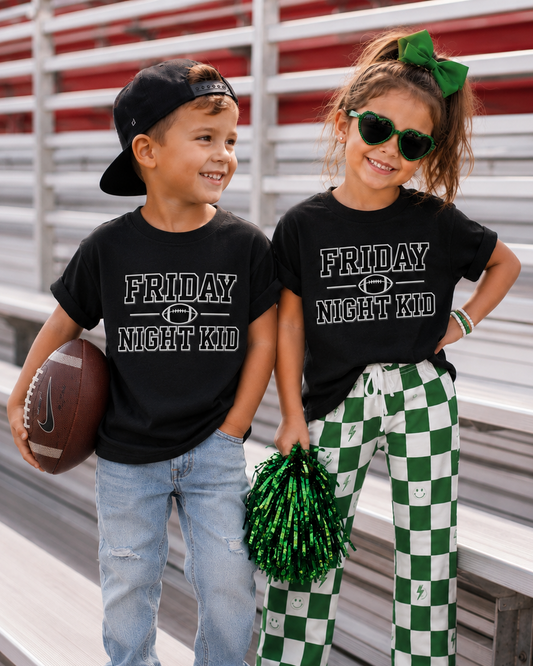 Two children standing on stadium bleachers wearing matching black graphic T-shirts that read 'FRIDAY NIGHT KID' in white text. The boy wears light blue jeans and a black cap, holding a football, while the girl wears green-and-white checkered pants, sunglasses, and a green hair bow, holding green pom-poms.